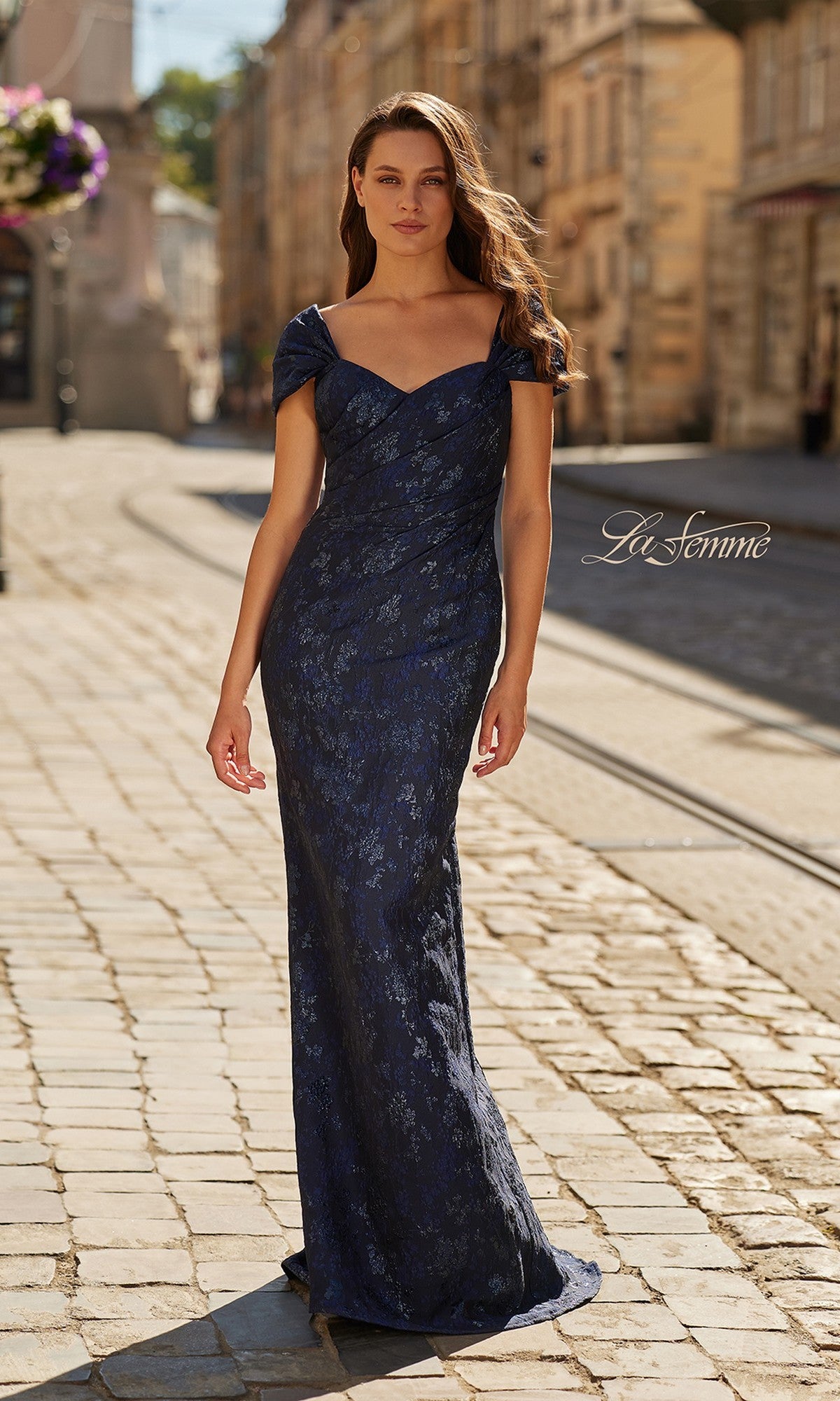 A woman in the navy Long Formal Dress: La Femme 33571 poses gracefully on a cobblestone street, her off-shoulder lace gown flowing elegantly in the daylight.